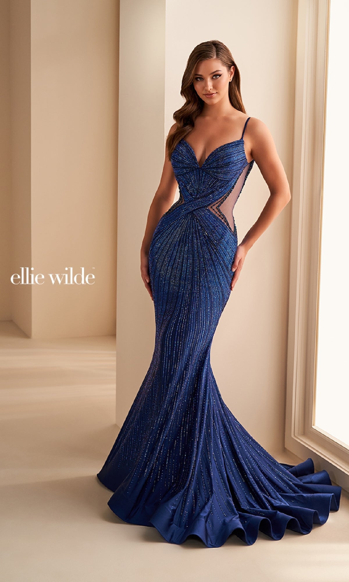 A woman stands by a window wearing the Sheer-Side Ellie Wilde Designer Prom Dress EW35001—a deep blue, beaded mermaid gown with thin straps, mesh side panels, a fitted silhouette, slight train, and striking details.